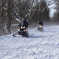 Two snowmobile riders on a snowy trail in the woods.